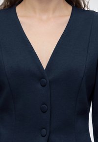 Gros plan d'une personne portant un vêtement bleu marine à col en V avec trois grands boutons recouverts de tissu assorti sur le devant.