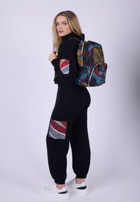 Kvinna i svart outfit med färgglada lappar bär en flerfärgad mönstrad ryggsäck och vita sneakers, står mot en enfärgad bakgrund.