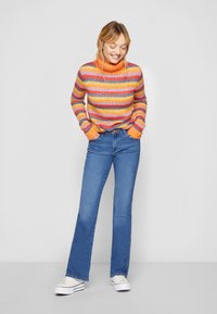 Strikket høyhalset genser med flerfargede horisontale striper i oransje, rosa og lilla; kombinert med blå svingjeans og sneakers.
