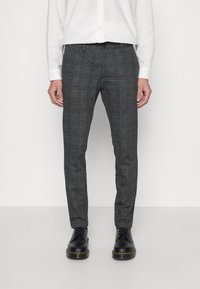 Pantalons à carreaux gris en tissu texturé, affichant une coupe slim et des passants de ceinture, associés à des chaussures noires et une chemise blanche à boutons.