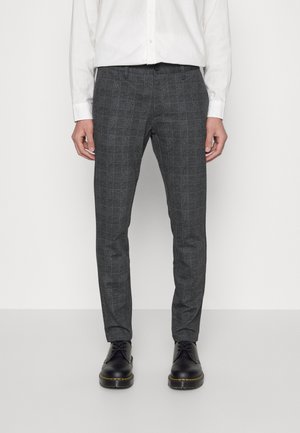 Pantalons à carreaux gris en tissu texturé, affichant une coupe slim et des passants de ceinture, associés à des chaussures noires et une chemise blanche à boutons.