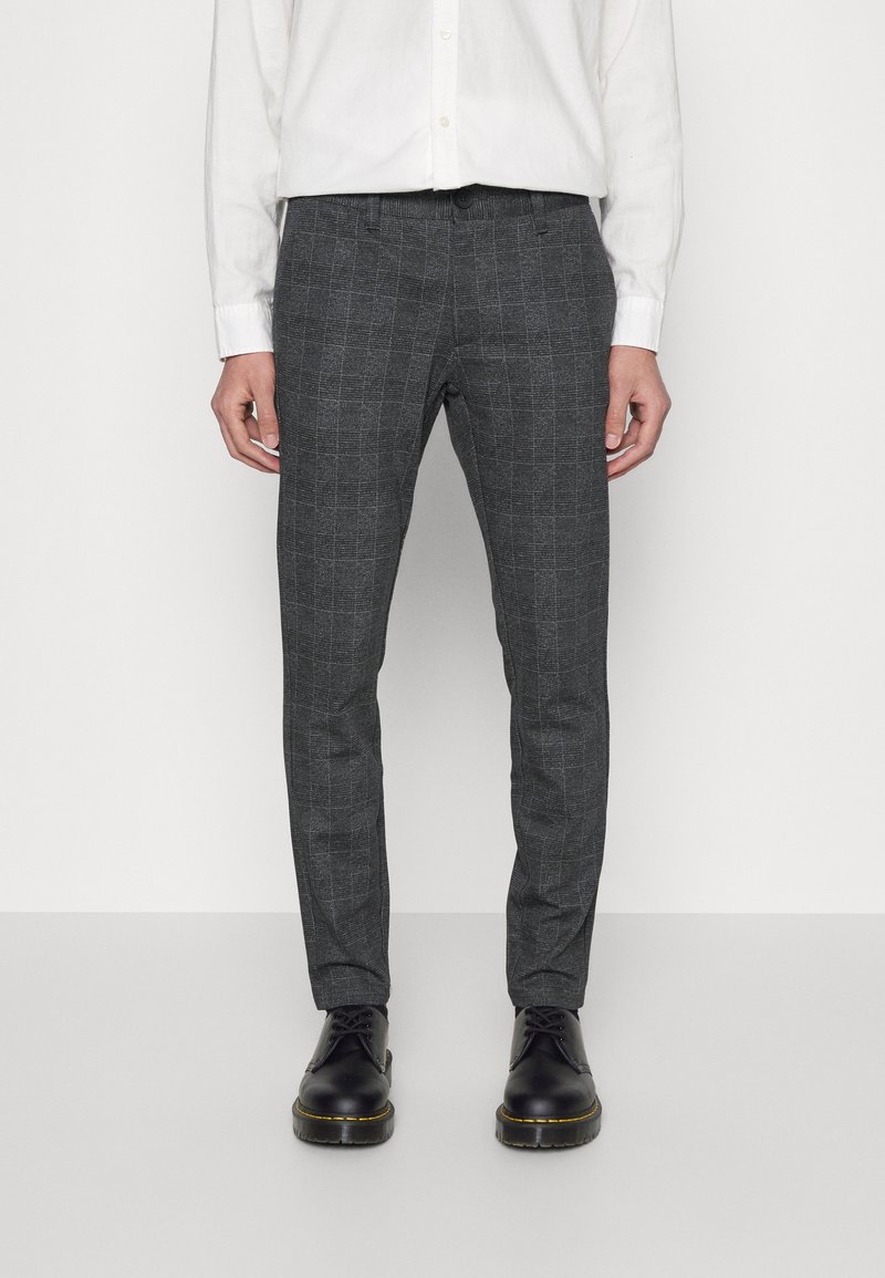 Pantalons à carreaux gris en tissu texturé, affichant une coupe slim et des passants de ceinture, associés à des chaussures noires et une chemise blanche à boutons.
