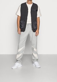 Černá prošívaná vesta s dvěma předními kapsami, spárovaná se šedými tepláky s bílými diagonálními akcenty a bílými teniskami.