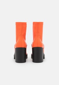 Botas de tobillo de cuero naranja con una textura suave, presentan un tacón grueso negro y costuras verticales en la parte trasera.