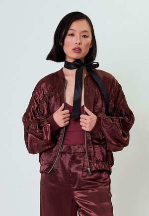 Burgundy satin bomber jacket with puffed sleeves and elastic cuffs, paired with a matching top and pants. Features a center zip and rounded collar.