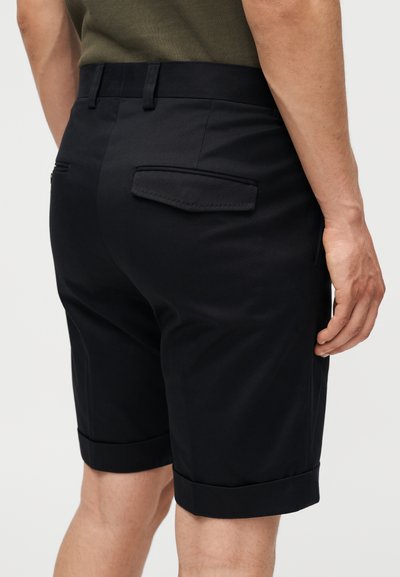 Άνδρας που φοράει μαύρα, ραμμένα στα μέτρα σορτς με γυρισμένα τελειώματα και πίσω τσέπες με καπάκια, συνδυασμένα με πουκάμισο ελιάς.