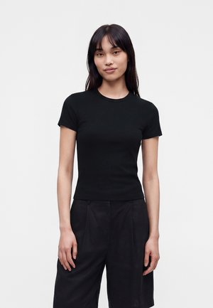 Donna in piedi contro uno sfondo semplice, indossa una t-shirt nera aderente a maniche corte e pantaloni neri larghi, rivolta in avanti con espressione neutra.