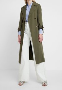 Oliwkowy dwurzędowy płaszcz trench z dużymi guzikami, podwiniętymi rękawami; w zestawieniu z jasnoniebieską koszulą i szerokimi białymi spodniami.