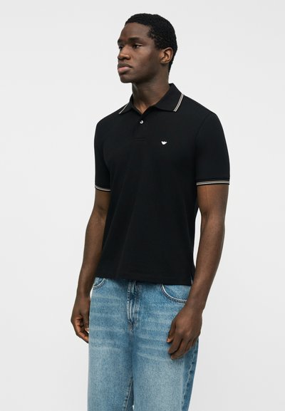 Άνδρας που φοράει μαύρο πόλο με κοντό μανίκι με λευκή ρίγα στα τελειώματα και ανοιχτό μπλε τζιν, στέκεται μπροστά σε απλό λευκό φόντο.