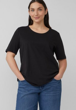 Vrouw staat met handen in de zakken, gekleed in een eenvoudige zwarte T-shirt met korte mouwen en blauwe jeans, tegen een grijze achtergrond.