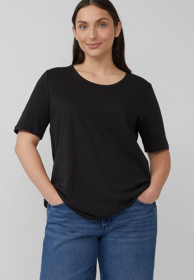 Eine Frau steht mit den Händen in den Taschen, trägt ein einfaches schwarzes kurzes Hemd und blaue Jeans vor einem grauen Hintergrund.