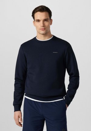 Hombre que lleva una sudadera azul marino con cuello redondo y pequeño logo de "Hackett" en el pecho y pantalones azul marino, de pie frente a un fondo claro y liso.