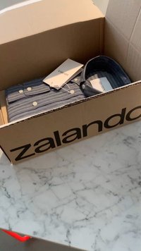 Pahvilaatikko, jossa on "Zalando"-etiketti, sisältää taitetun sinivalkoraitaisen paidan, jonka napit ja hintaletkut ovat näkyvissä, lepäämässä marmoripinnalla.