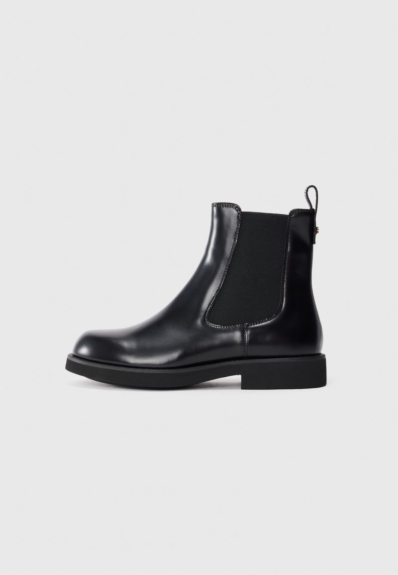 Stivale Chelsea in pelle nera con pannello laterale elasticizzato, linguetta sul tallone, suola bassa a blocco, esposto su sfondo bianco.