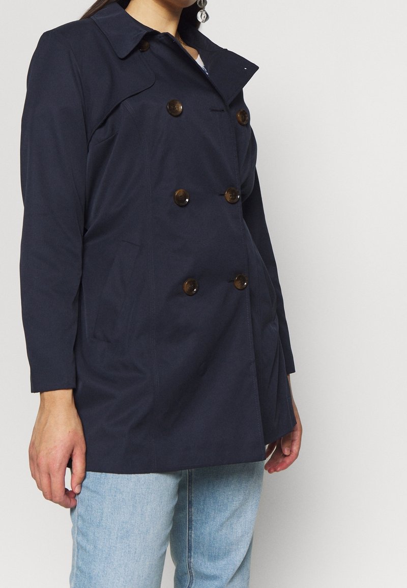 Femme portant un manteau croisé bleu marine foncé avec de gros boutons et un jean bleu clair sur un fond clair uni.