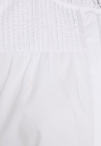Tessuto di cotone bianco con pieghe verticali nella parte superiore, texture liscia e design fluido con dettaglio arricciato in vita.