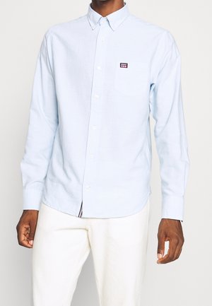 Homme portant une chemise bleu clair boutonnée avec un logo sur la poche poitrine, associée à un pantalon blanc, debout devant un fond uni.