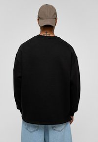 Zwarte oversized sweatshirt van katoen, met een ronde hals, verlaagde schouders en rib gebreide manchetten. De persoon draagt een grijze baseballcap.