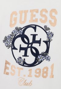 Balts auduma T-krekls ar tumši zilu izšūtu logotipu ar ziedu akcentiem un metalizētu zelta tekstu, kas lasāms "GUESS," "EST. 1981," un "Club."