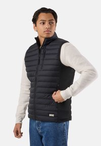 Giovane uomo con indosso un gilet nero trapuntato senza maniche sopra una camicia a maniche lunghe beige chiaro e jeans blu, che guarda di lato con una mano sulla cerniera del gilet.