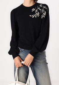 Suéter negro de manga larga abullonada, con detalles de hojas bordadas en blanco en los hombros; combinado con jeans de mezclilla azules.