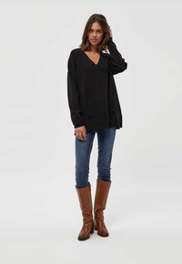 Črni oversize pulover z V-izrezom, dolgimi rokavi in rebrastimi manšetami, v kombinaciji z oprijetimi modrimi kavbojkami in rjavimi škornji do kolen.