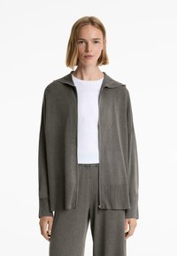 Grå zip-up cardigan med huva, ribbad textur, vida ärmar och lös passform, i kombination med en vit topp och matchande grå byxor.