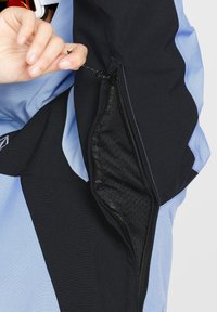 Zwarte en lichtblauwe jas met een ritszak, voorzien van een mesh voering. Het gladde materiaal en de contrasterende kleurpanelen zijn zichtbaar.