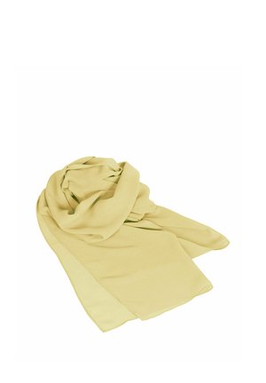 Kvinde står iført en skulderfri, flydende lysegul kjole med et matchende halstørklæde og et guldarmbånd på hendes højre håndled.