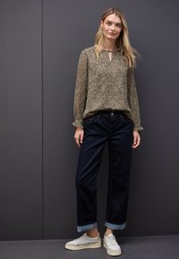 Jonge vrouw met lang blond haar, gekleed in een beige blouse met stippen met lange mouwen, donkere opgerolde jeans en witte sneakers, staand tegen een grijze muur.