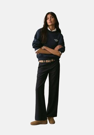 Marineblauwe oversized sweatshirt met logo, witte kraag, hooggewaisted gestreepte broek en tan suede schoenen. Eenvoudig, ontspannen ontwerp.