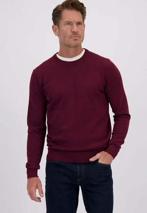 Pull en tricot bourgogne avec un col rond et des poignets côtelés, associé à un jean en denim foncé. Texture douce, design ajusté.