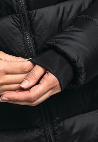Schwarze Pufferjacke mit gerippten Bündchen. Der Stoff hat eine dezente Gitterstruktur, und der Reißverschluss verläuft vertikal an der Vorderseite. Die Hände greifen das Bündchen.