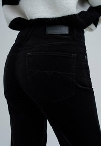 Gros plan sur une personne portant un jean en velours côtelé noir étiqueté "Salsa Jeans" avec un pull rayé noir et blanc rentré.