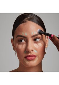 Visage d'un mannequin avec différentes couleurs de crème appliquées par sections : beige clair, vert et pêche. Un pinceau est utilisé pour estomper. Texture de peau lisse.
