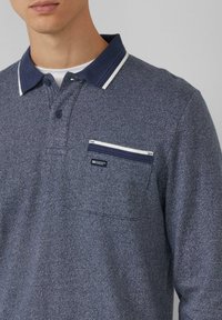 Granatowa koszulka polo z długim rękawem o fakturowanym wzorze, kołnierzyk w białe paski oraz kieszonka na piersi z ozdobnym zamkiem.