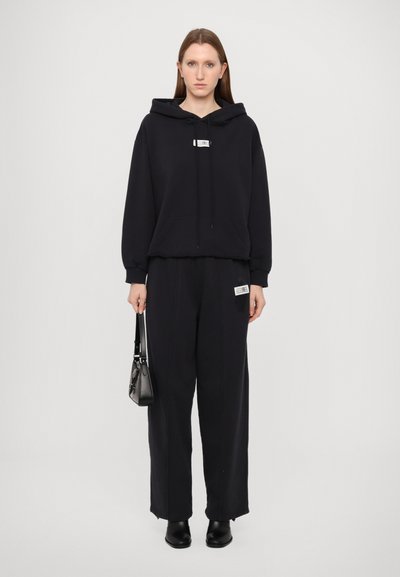 Svart hoodie med justerbara snoddar och känguruficka, kombinerad med matchande lösa träningsbyxor. Litet logopatch och svarta ankelboots.