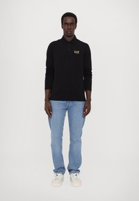 Juodas ilgomis rankovėmis polo marškinėliai su geltonu logo, derinami su šviesiai mėlynomis džinsais ir baltais sportbačiais, prieš paprastą foną.