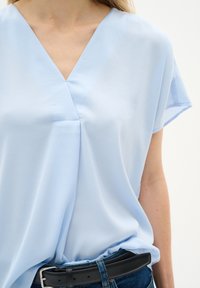 Blusa azzurra chiara a maniche corte con scollatura a V, realizzata in tessuto morbido e con una vestibilità rilassata. Accompagnata da una cintura nera sopra un paio di jeans.