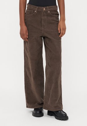 Persoon draagt bruine wijde ribfluwelen broek met voorzakken en knoopsluiting, gecombineerd met zwarte chunky schoenen en een zwart shirt.