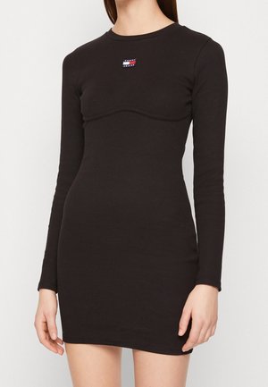 Femme portant une mini-robe noire ajustée à manches longues avec un petit logo rectangulaire Tommy Jeans sur la poitrine.