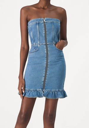 Strapless blue denim mini dress with front zipper, pockets, and ruffled hem, worn by a person with one hand in pocket.
