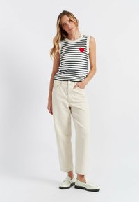 Top senza maniche a righe con una toppa a forma di cuore rossa, abbinato a pantaloni a gamba larga color crema e scarpe slip-on bianche con dettagli neri.
