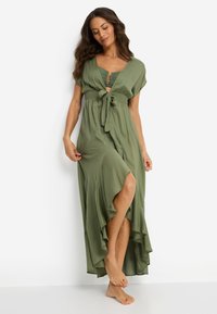 Μάξι φόρεμα ελιάς με κοντά μανίκια, δέσιμο στη μέση και βολάν στο τελείωμα. Διαθέτει βαθύ V λαιμόκοψη και σκίσιμο μέχρι το μέσο του μηρού. Ελαφρύ ύφασμα.