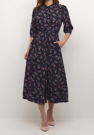 Femme portant une robe midi bleu marine avec un motif floral rose et violet, manches trois-quarts, devant boutonné et ceinture nouée à la taille.
