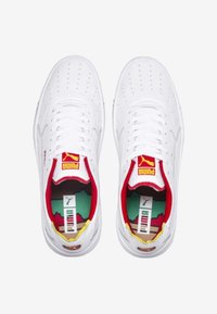 Ett par vita Puma-sneakers med röda och gula accenter, perforerat tåsarea och märkta innersulor med en färgglad design.