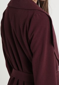 Bordowy płaszcz kopertowy z dużym kołnierzem, długimi rękawami, fakturowanym materiałem i paskiem w talii, z plecionymi detalami.