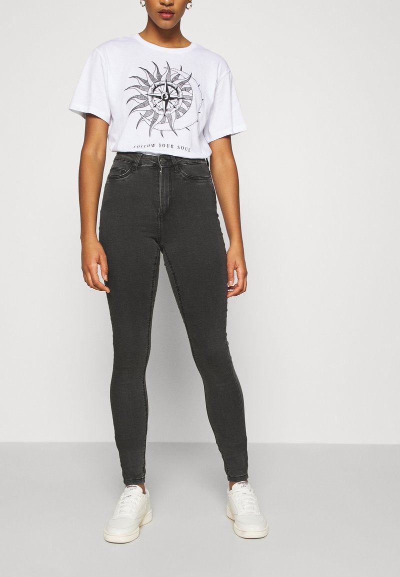 Camiseta gráfica blanca con un diseño circular del sol y texto arriba, combinada con jeans ajustados gris oscuro y zapatillas blancas.