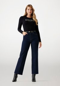 Maglietta nera a maniche lunghe con logo "GUESS", abbinata a jeans a gamba larga blu scuro e stivaletti neri con tacco; tessuto morbido e design elegante.