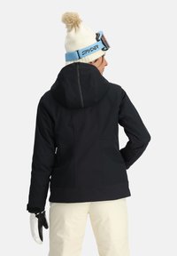 Schwarze Skijacke mit Kapuze, ausgestattet mit strukturierten Einsätzen. Trägt dazu eine helle Hose und weiße Handschuhe. Eine helle Mütze mit Pompon sitzt auf dem Kopf.